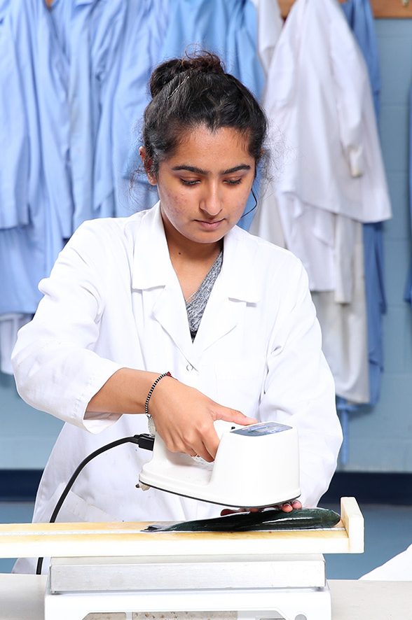 Two students and an instructor in lab coats working at a desk. One student types on a laptop, while the other uses an instrument to take a reading of a fish while the instructor looks on. There is a row of labcoats hanging from hooks in the background, with posters of fish above them.