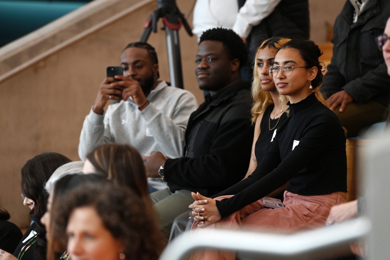 A group of diverse attendees sit on bleachers indoors at an event while one records on his phone.