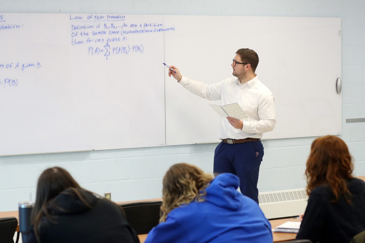 Male class instructor pointing at blue writing on a whiteboard. Students sit in the foreground.