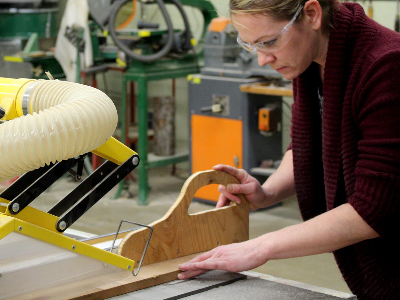 Student in Tech Ed woodworking shop operating a table saw with safety glasses and a safety push stick.