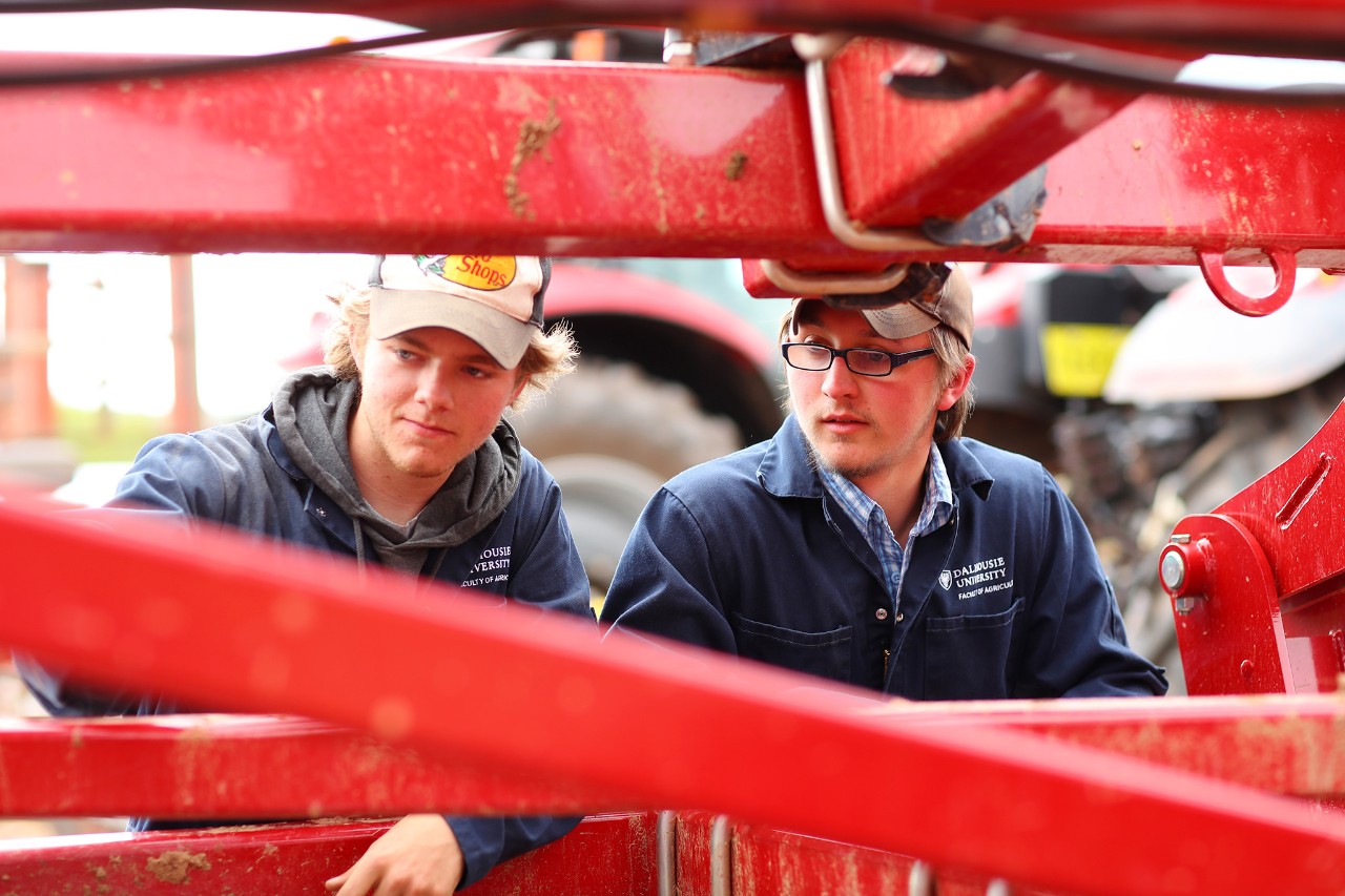 Two students wearing blue coveralls and baseball caps looking at a red tractor attachment.