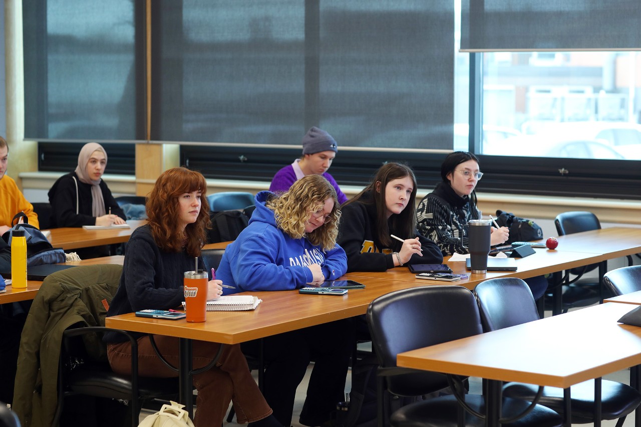 A group of students sit at rows of long wooden tables with notebooks or tablets in front of them. 
