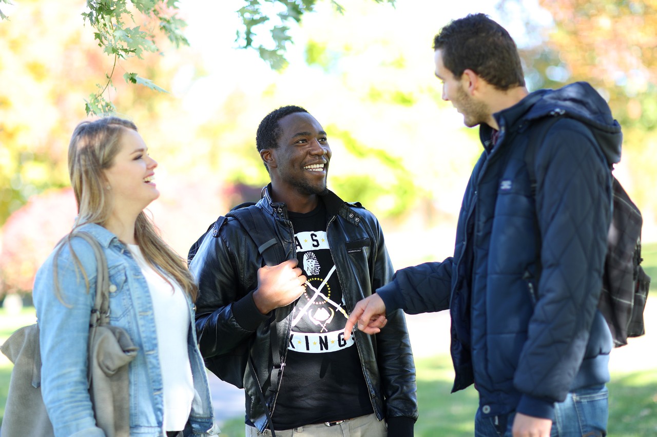 Three students stand chatting together in a group with leafy trees in the background.