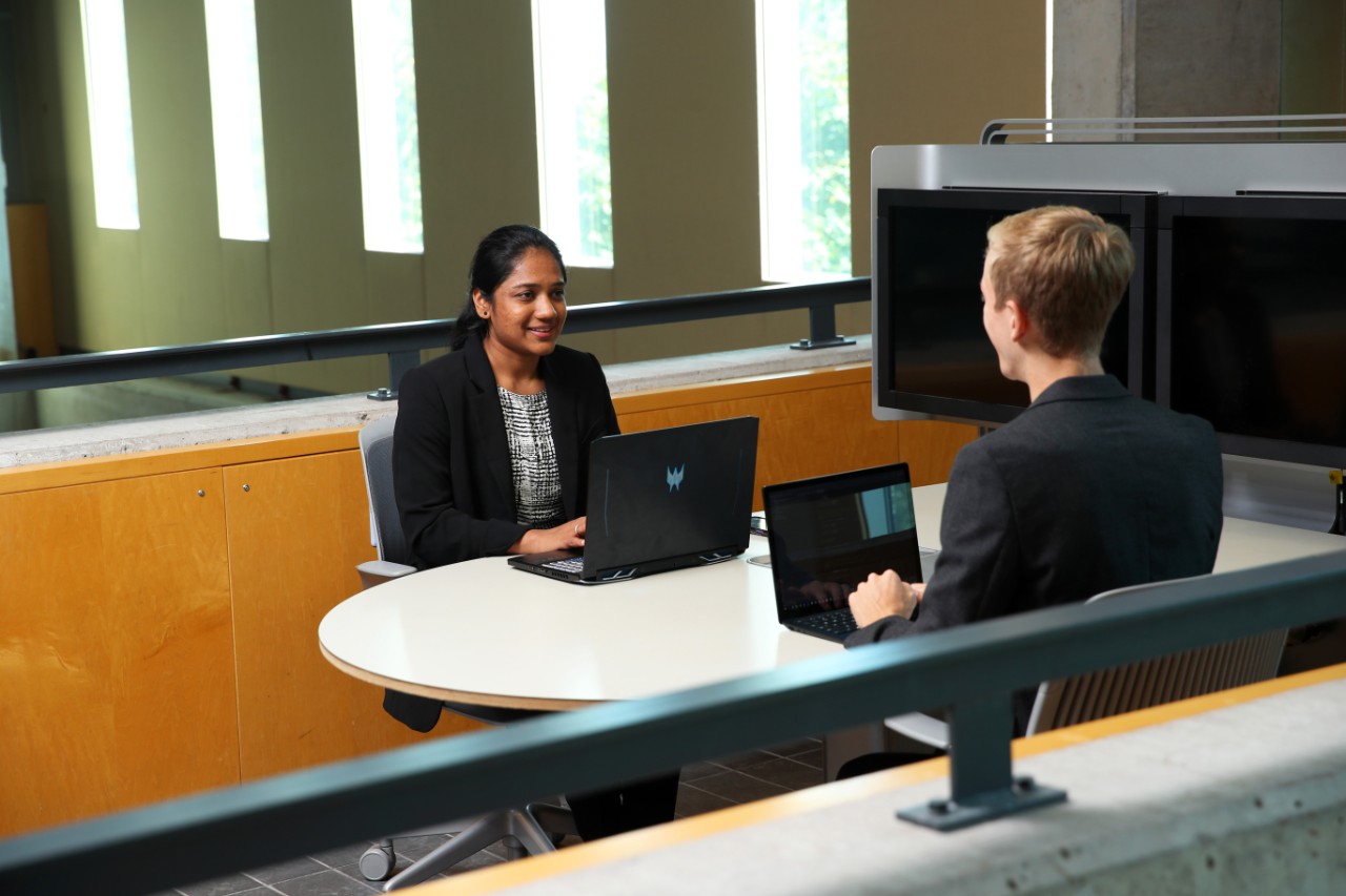 two students in business attire with laptops sit across from each other at a desk