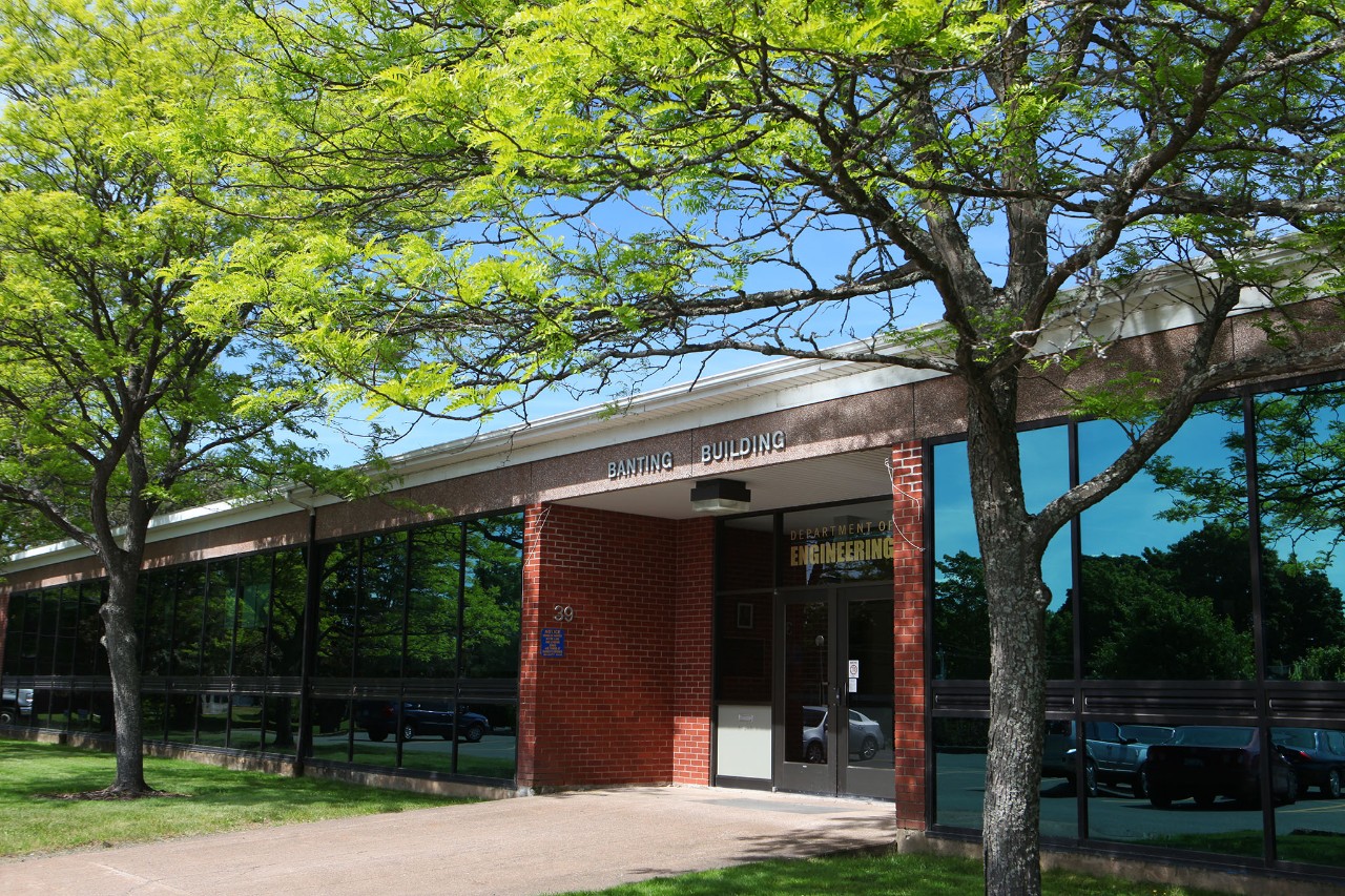 Entrance of low red brick building flanked by floor to ceiling window. The building is has a row of leafy green trees in front.