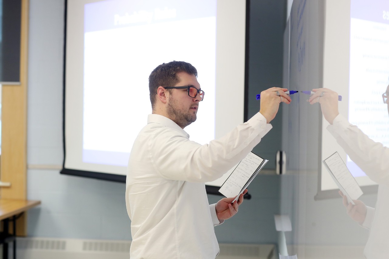 Male class instructor wearing a white shirt and black framed glasses writing lesson notes on a whiteboard.
