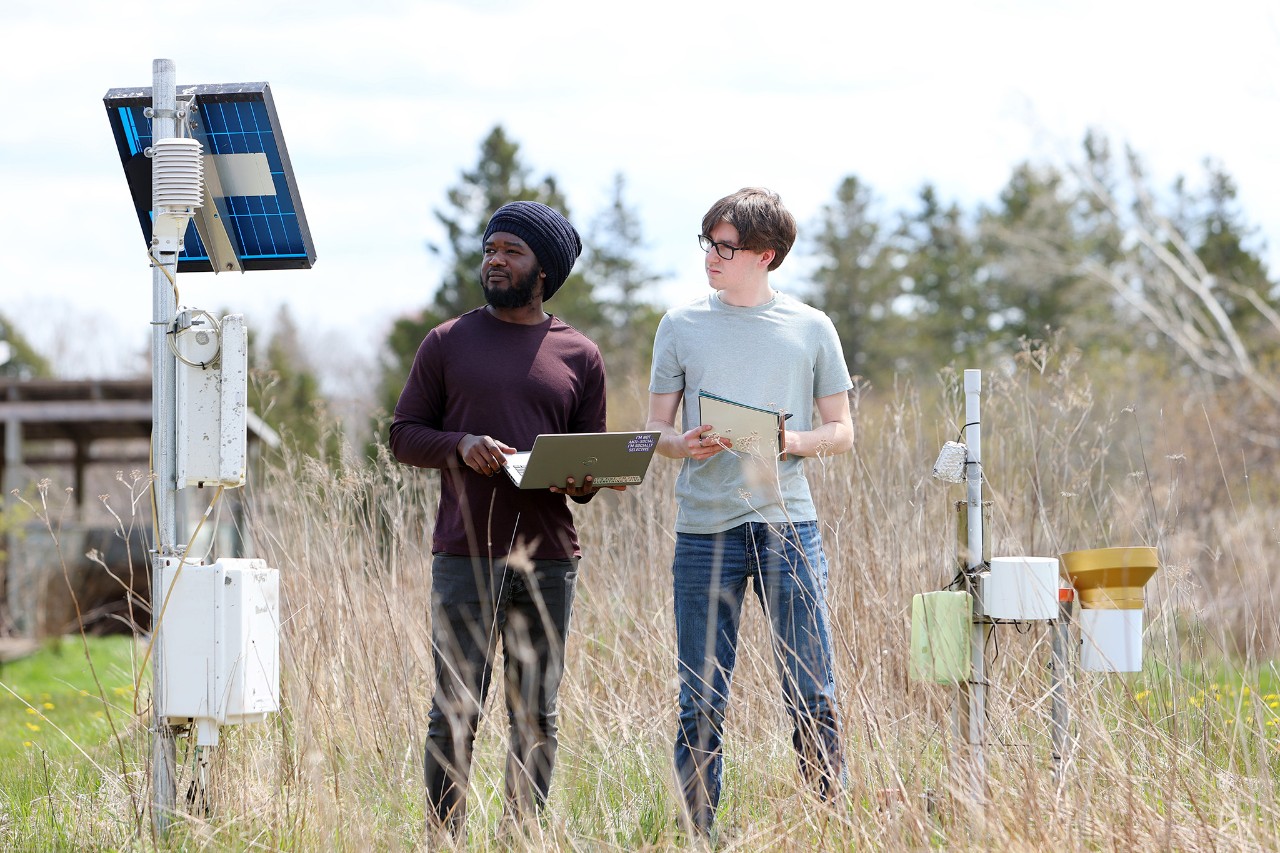 Two students standing in long grass in a field with engineering equipment around them. One student is holding a laptop, the other is taking notes on a pad.