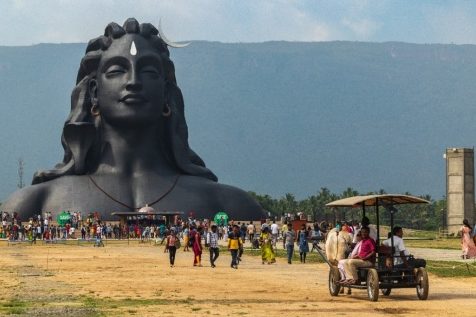 A large back sculpture depicting the head and shoulders of a figure with long hair rises out of the ground. People and wagons are in the foreground, and mountains can be seen in the background.