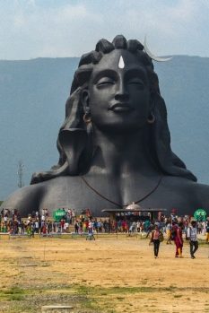 A large back sculpture depicting the head and shoulders of a figure with long hair rises out of the ground. People and wagons are in the foreground, and mountains can be seen in the background.