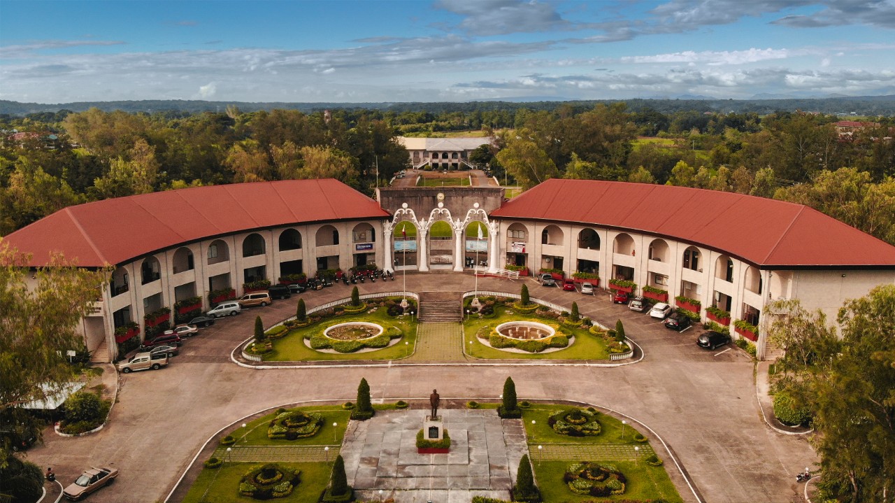 Curved beige stone building with red roof, surrounded by formal gardens.