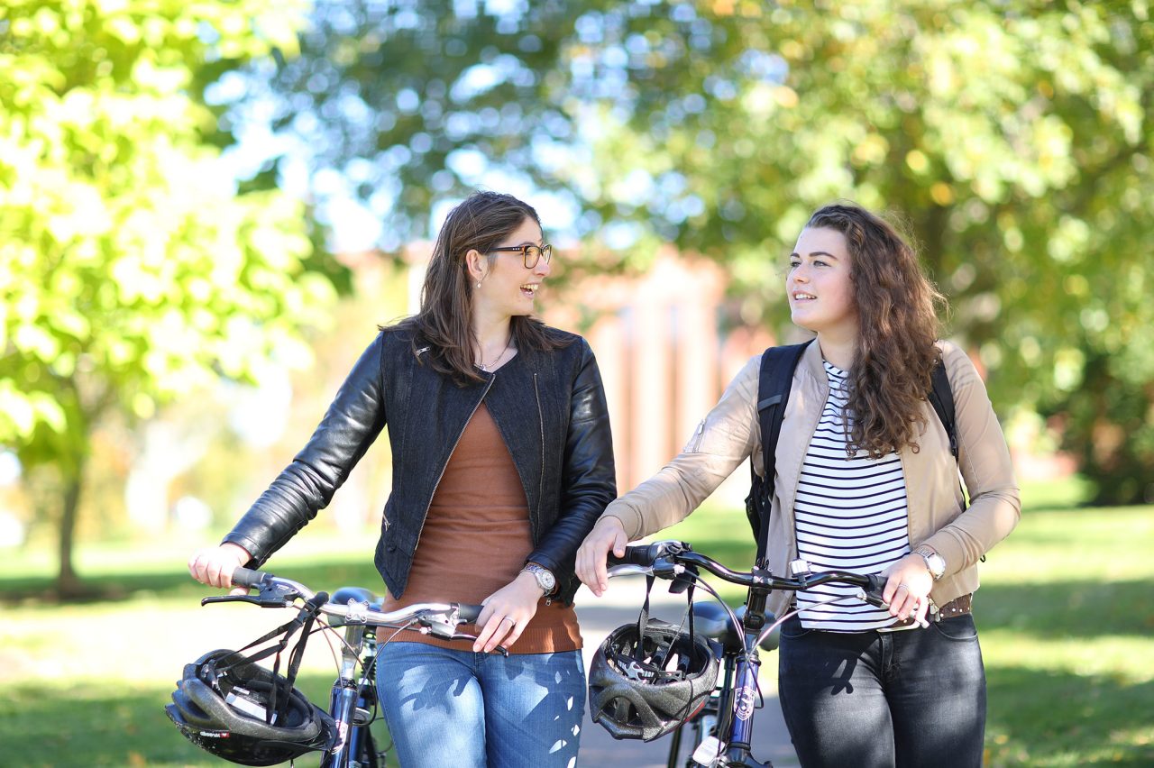 Two students walking their bikes down a path with green, leafy trees in the background. They are dressed casually, and their bike helmets hang from their handlebars.