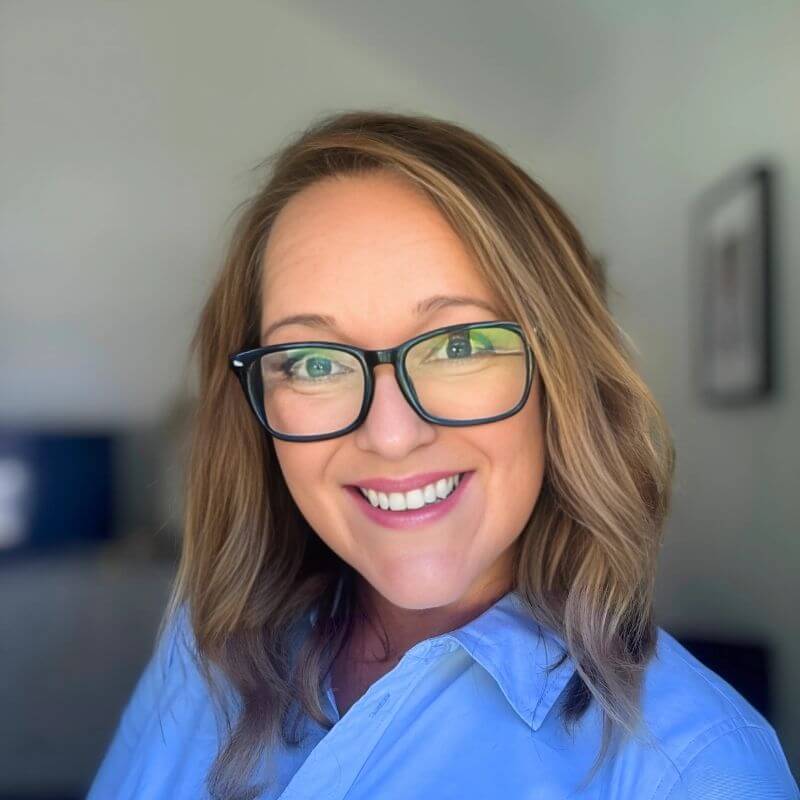  A smiling woman with shoulder-length blonde hair and glasses wearing a light blue button-down shirt.