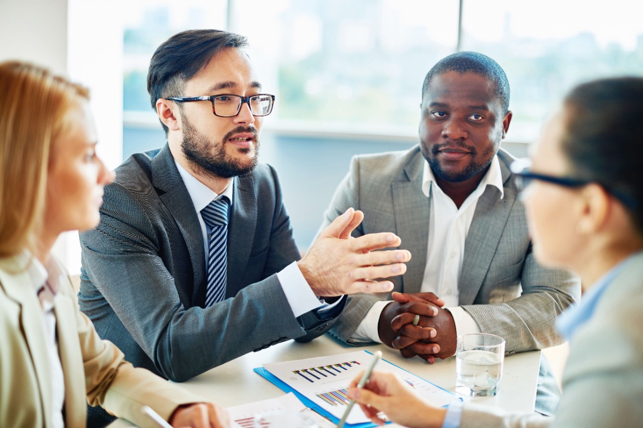 A group of diverse business people sitting around a table listening to a man who is speaking. 