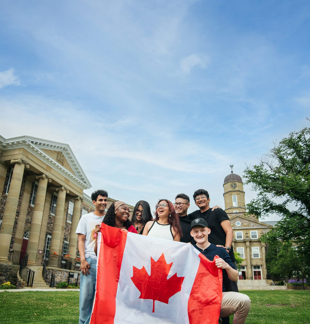 Seven students laugh while holding a Canada flag, posing for a photo in front of Dalhousie's Henry Hicks building.