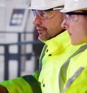 Two engineers wearing safety jackets and white hard hats sit at a desk looking at two computer monitors.