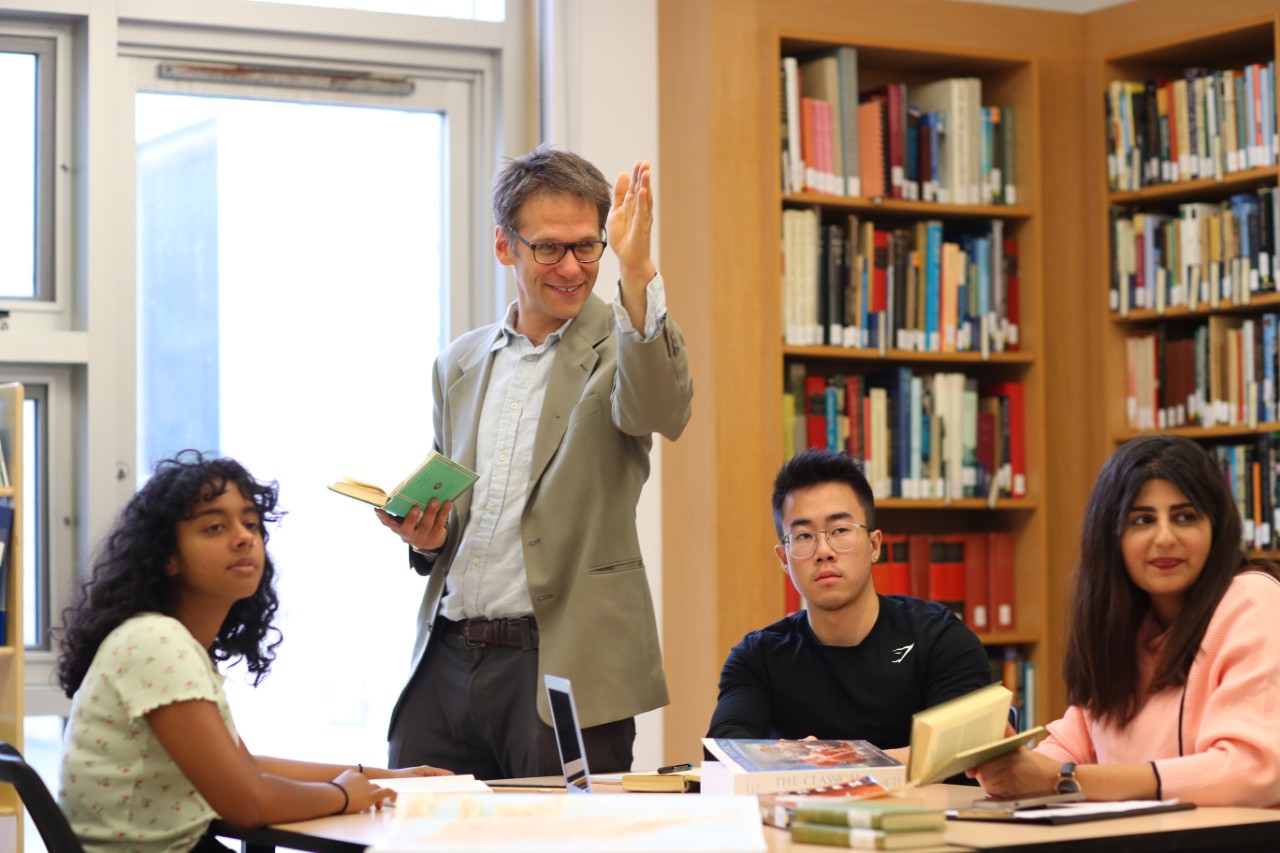 A male professor standing up and speaking to a small group of students who are seated at a table.