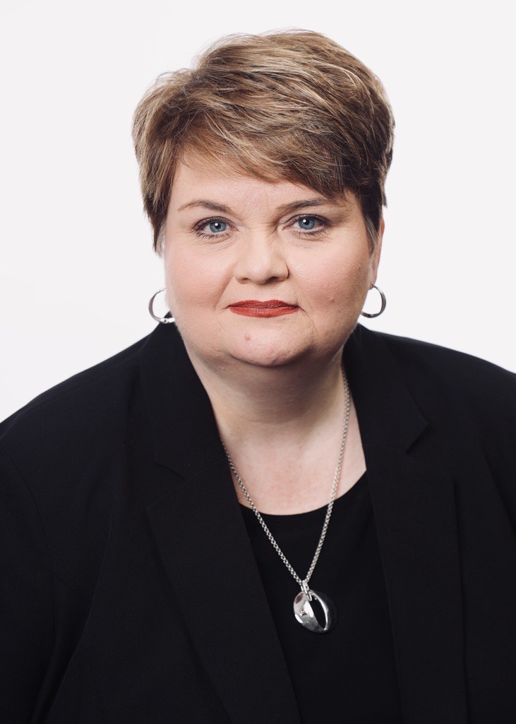 Woman with short light brown hair wearing a black blazer and silver pendant.