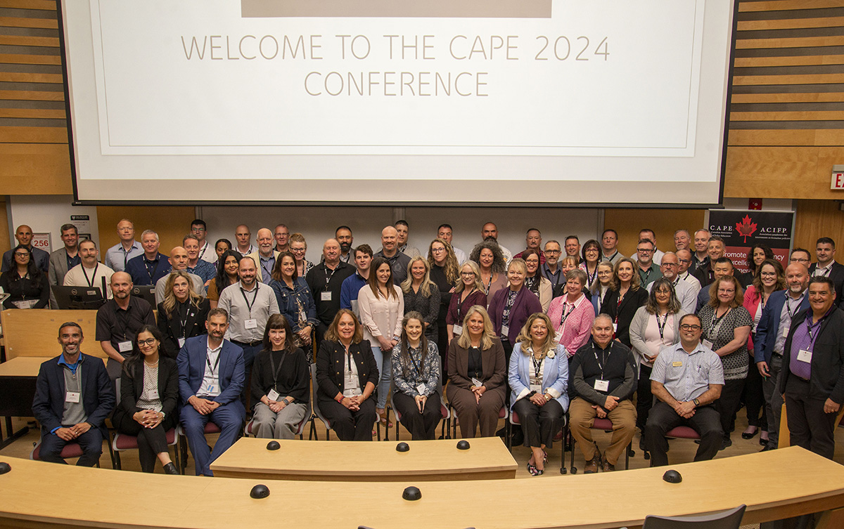 A group of individuals sit in a large classroom with a screen reading "Welcome to the 2024 CAPE Conference" behind them.