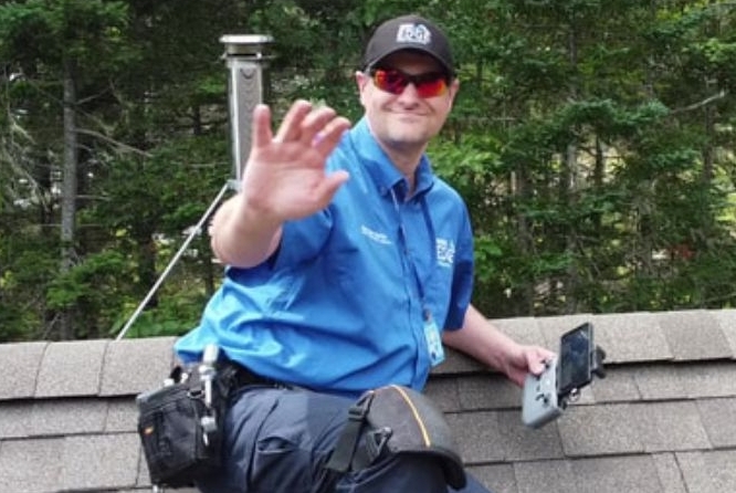 A man wearing sunglasses waving to the camera and safely working on a roof.