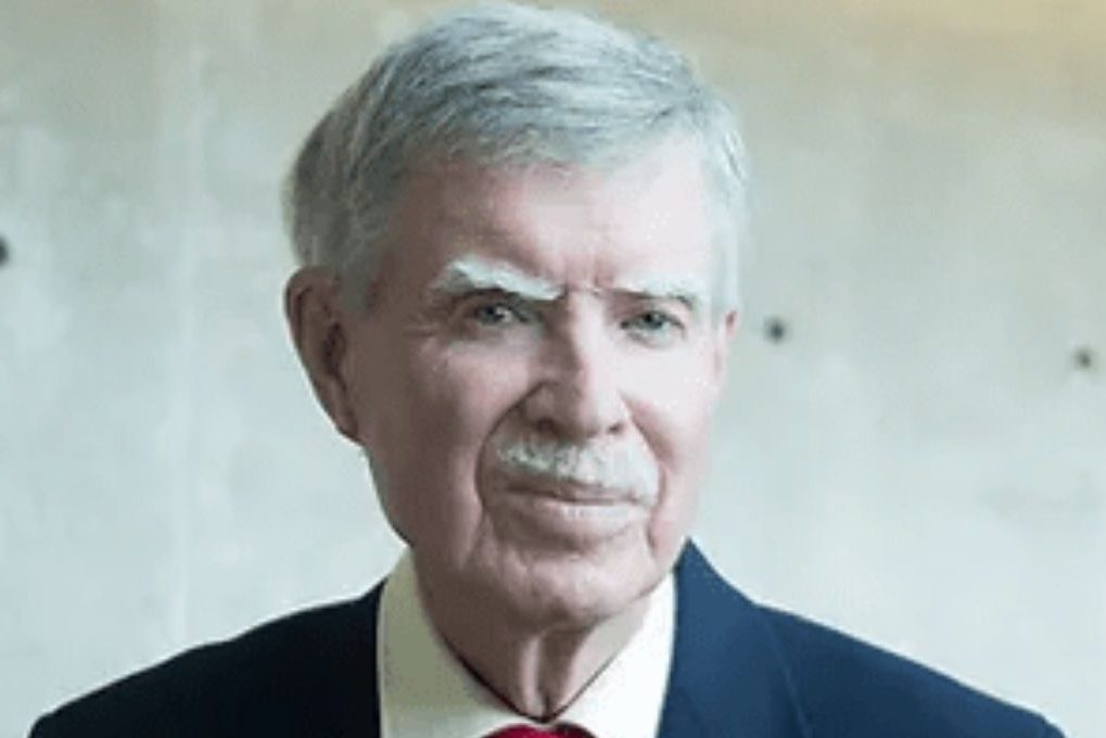 Headshot of a man with white hair wearing a blue suit and a red tie.