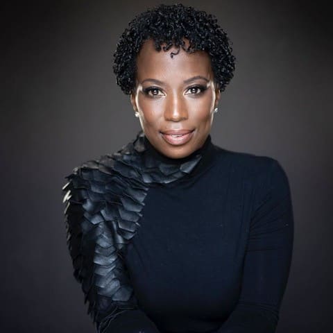 A studio portrait of a woman with short, tightly curled hair, wearing a black high-neck top with textured, layered detailing on one shoulder.