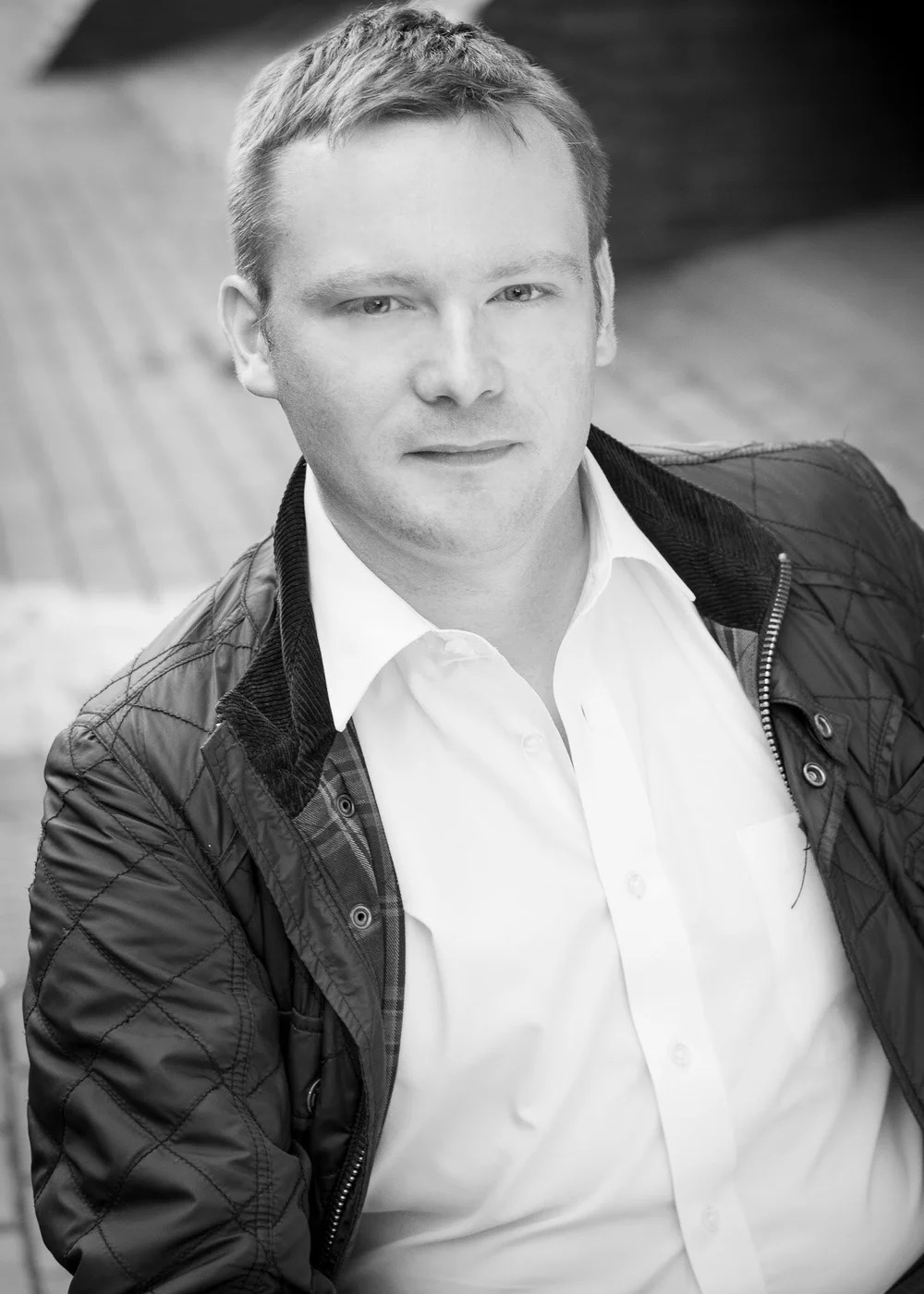 A black and white headshot of a man with short, light-colored hair, wearing a white button-down shirt under a quilted dark jacket, looking directly forward with a neutral expression.