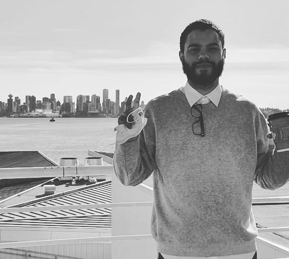 A black and white photo of Jason Zinn standing on an outdoor balcony with a city skyline and water in the background.