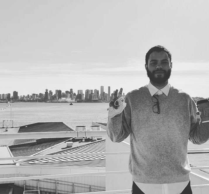 A black and white photo shows a man with a beard standing on a balcony overlooking a city skyline and water, holding a face mask and a coffee cup.