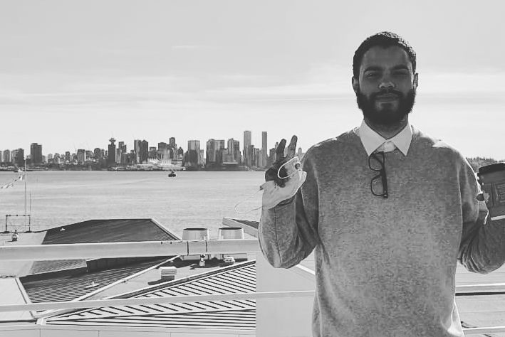 A black and white photo shows a man with a beard standing on a balcony overlooking a city skyline and water, holding a face mask and a coffee cup.
