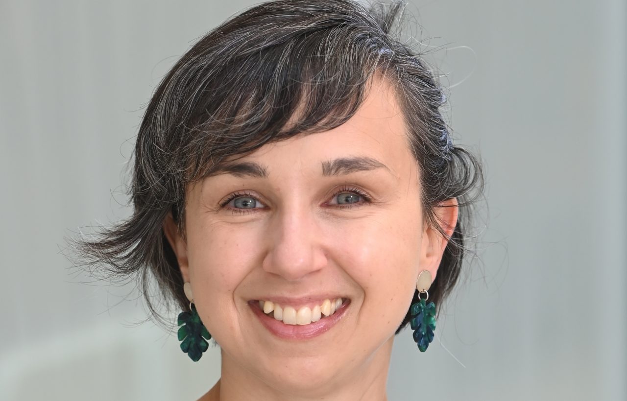 A smiling woman with short dark hair and green leaf-shaped earrings wears a dark green sweater in a professional headshot.