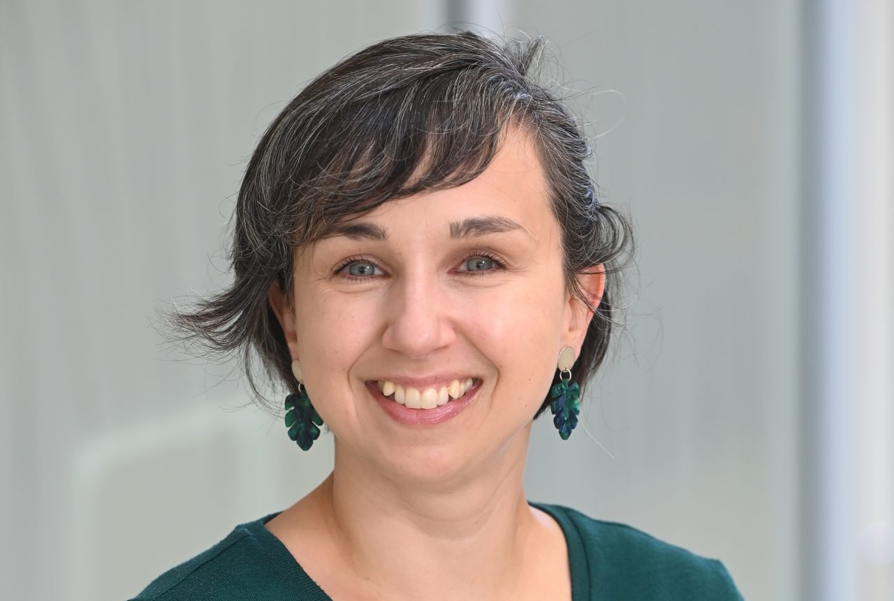 A smiling woman with short dark hair and green leaf-shaped earrings wears a dark green sweater in a professional headshot.