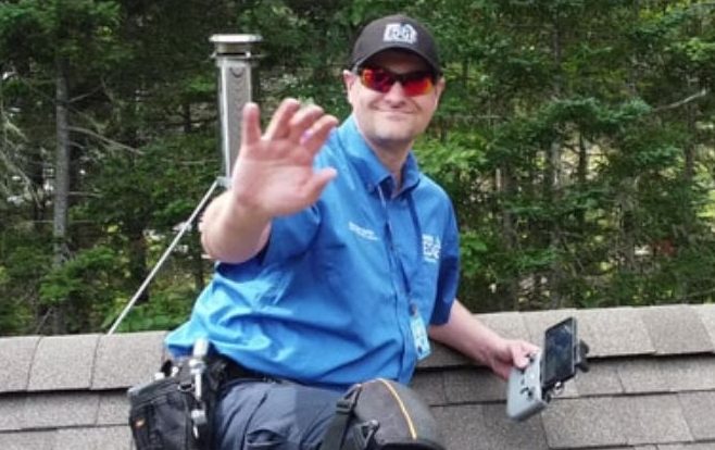 A man wearing sunglasses waving to the camera and safely working on a roof.