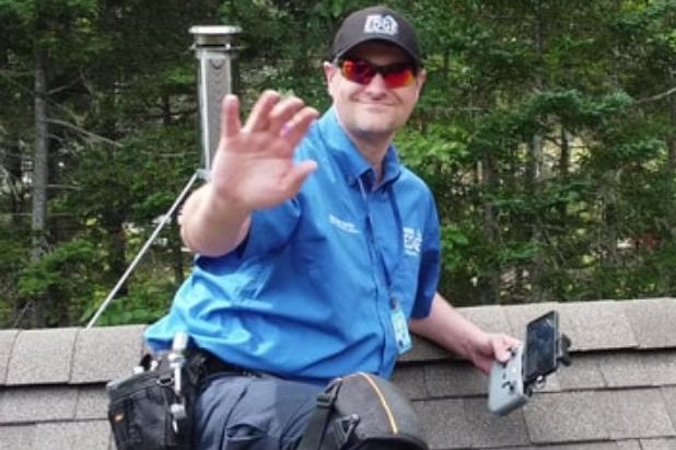 A man wearing sunglasses waving to the camera and safely working on a roof.