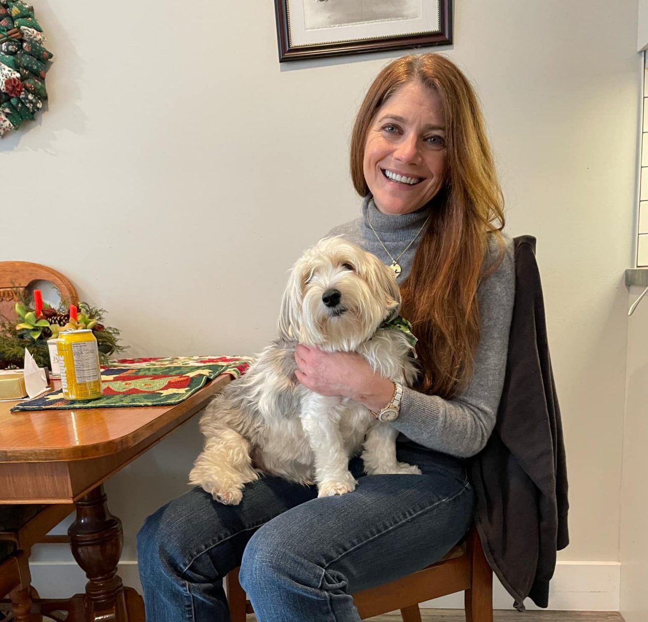  A smiling woman with long reddish-brown hair sits at a wooden table holding a fluffy white and grey dog on her lap.