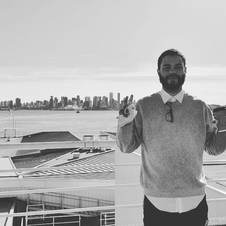 A black and white photo of Jason Zinn standing on an outdoor balcony with a city skyline and water in the background.