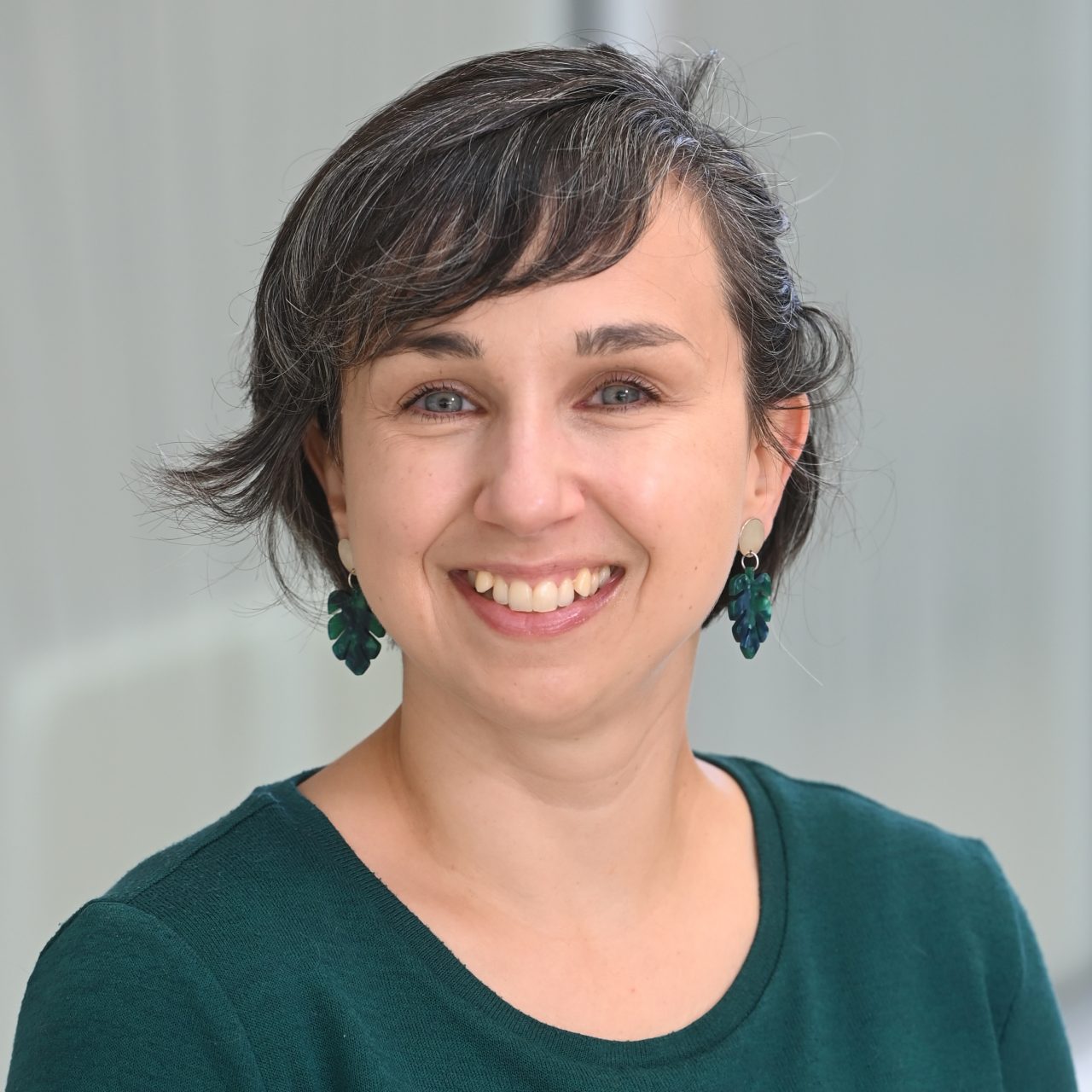 A professional headshot of a woman with short, dark hair and a warm smile, wearing a dark green long-sleeved shirt.