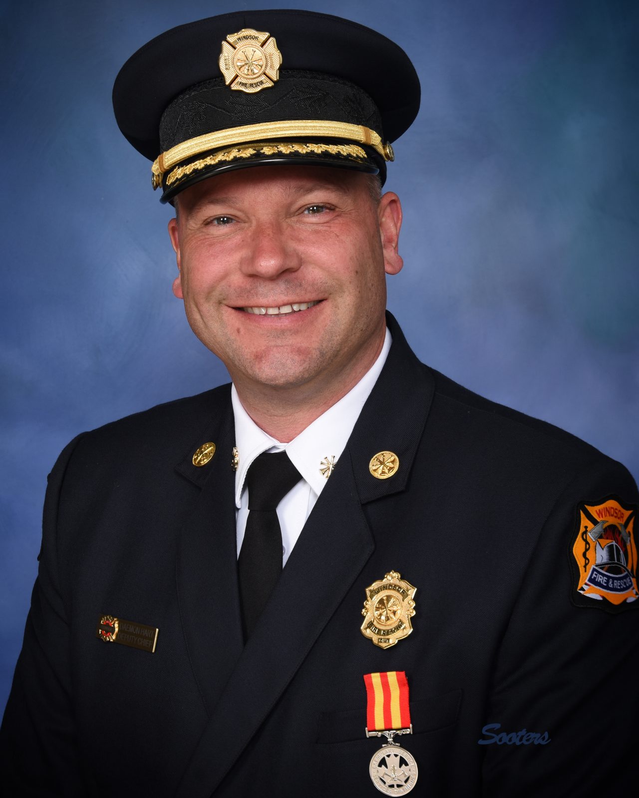Professional portrait of Daemon Hart, Deputy Fire Chief of Operations, in a formal navy blue Windsor Fire & Rescue dress uniform against a blue background.