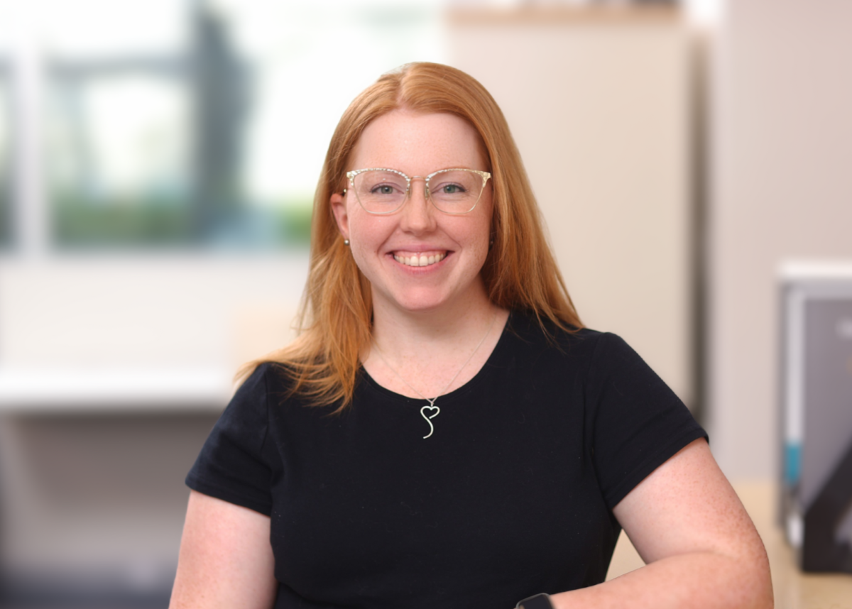 A professional headshot of a smiling woman with long red hair and gold-rimmed glasses, wearing a black t-shirt