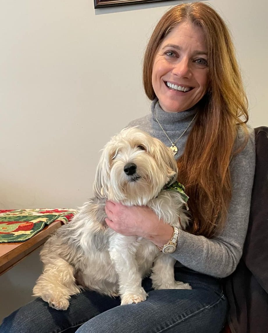 A smiling woman with long reddish-brown hair sits at a wooden table holding a fluffy white and grey dog on her lap.