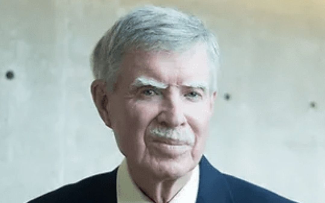 Headshot of a man with white hair wearing a blue suit and a red tie.