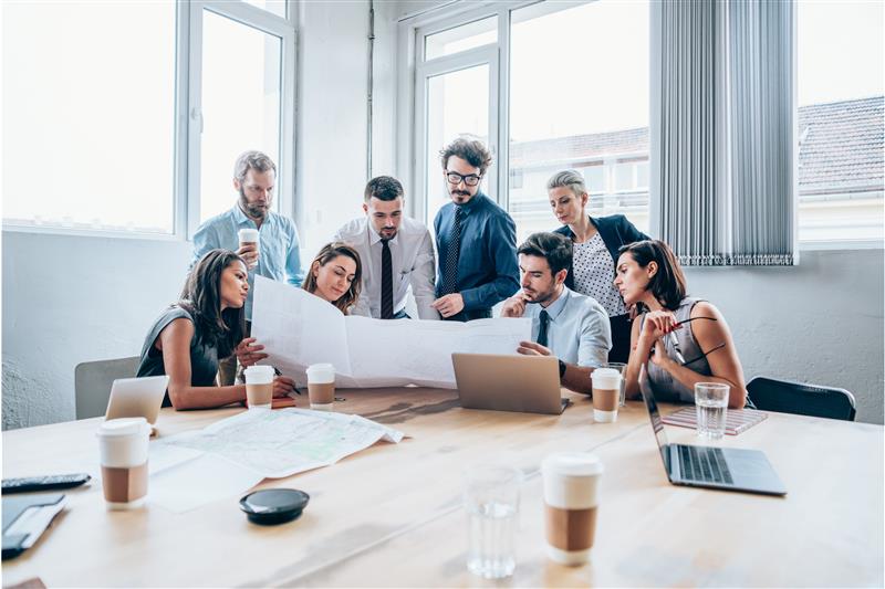 A group of diverse people sitting and standing around a table discussing details on a large sheet of paper.