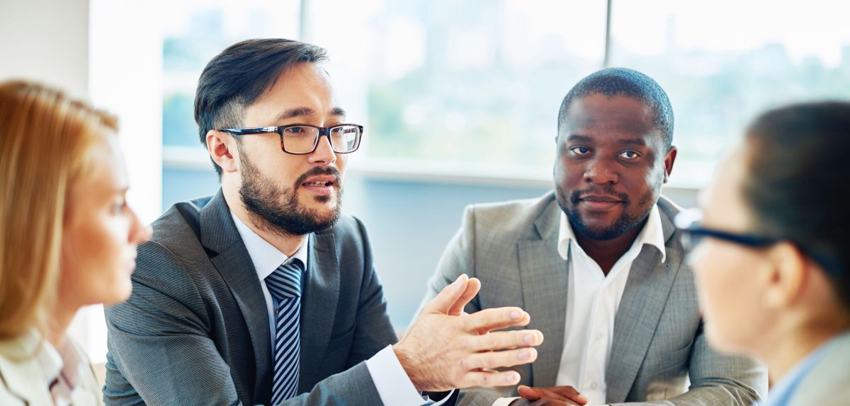 A group of diverse business people sitting around a table listening to a man who is speaking.