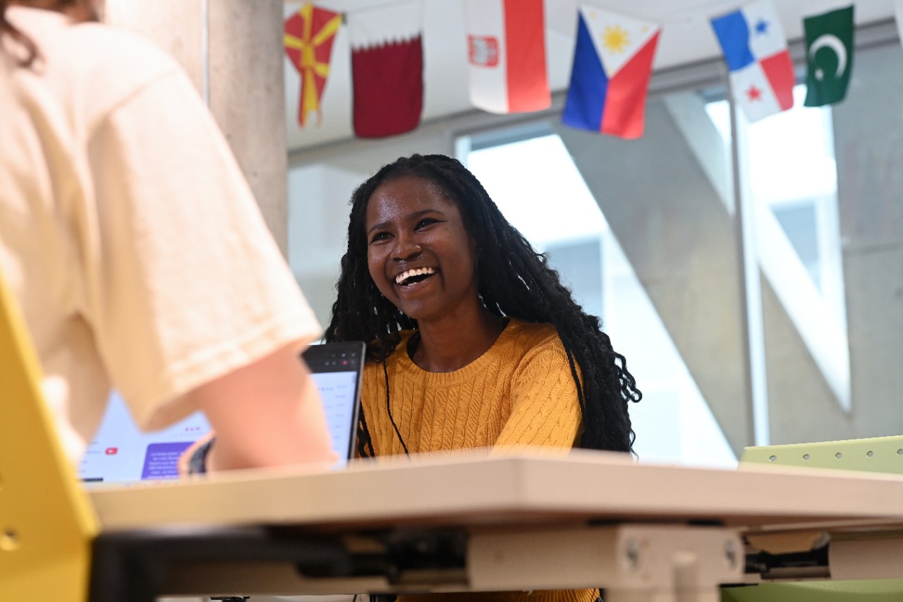 A student in a yellow sweater laughing, while sitting at a table in the International Centre, with flags of the world in the background.