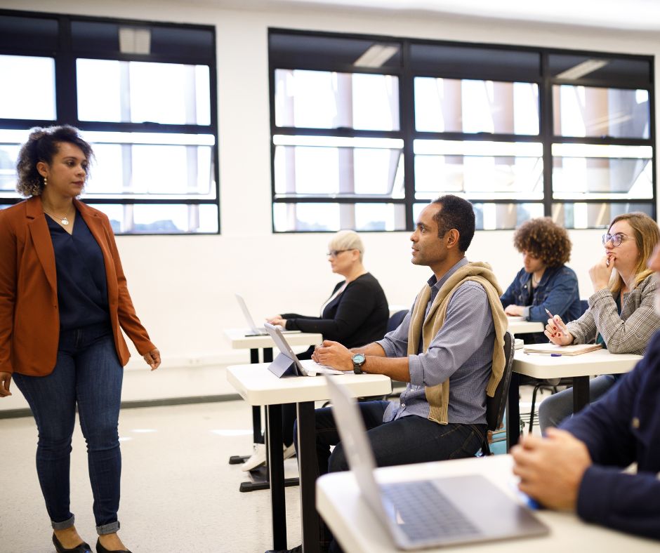 A woman standing in-front of a group of diverse adult students in a classroom.