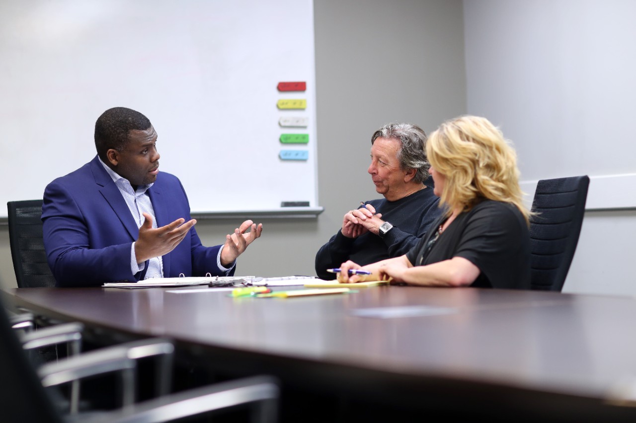 three people sitting in a conference room where a black man is speaking to another woman and man.