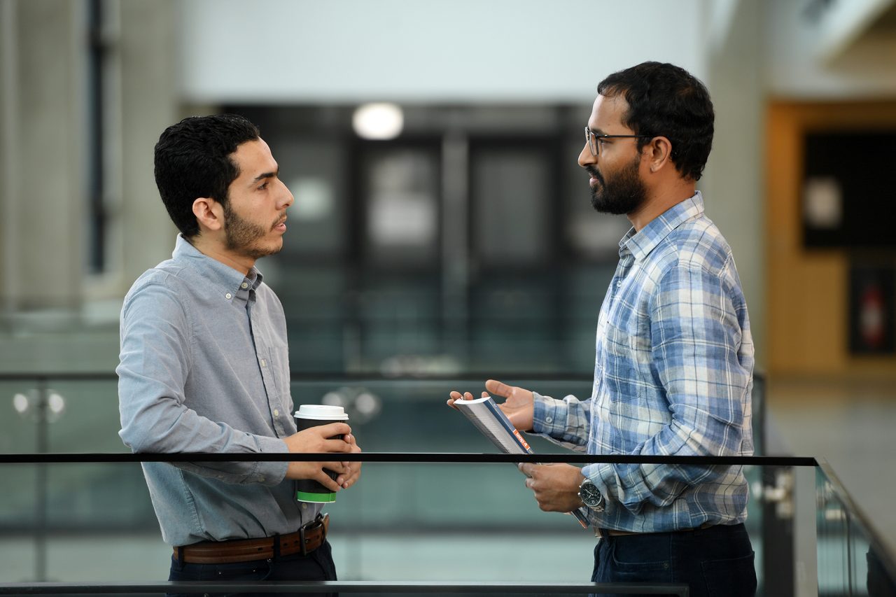 Two students shown from the side. One is carrying a coffee cup, and the other has a notebook.