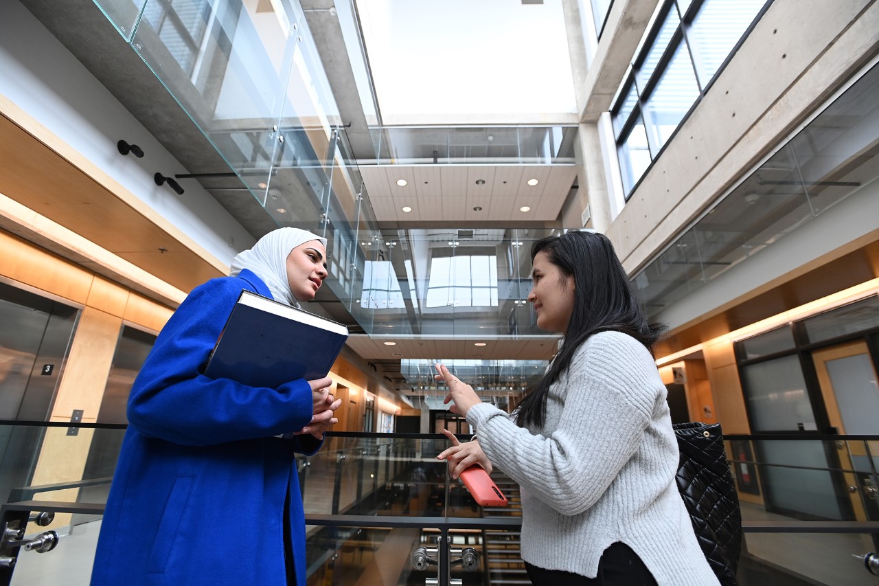 Two female students chat while leaning on a stair rail in a modern hallway with skylight.
