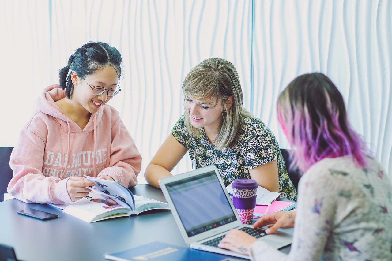 3 students collaborating at a table with their notes. One works on a laptop, and the other two flip through a textbook together.