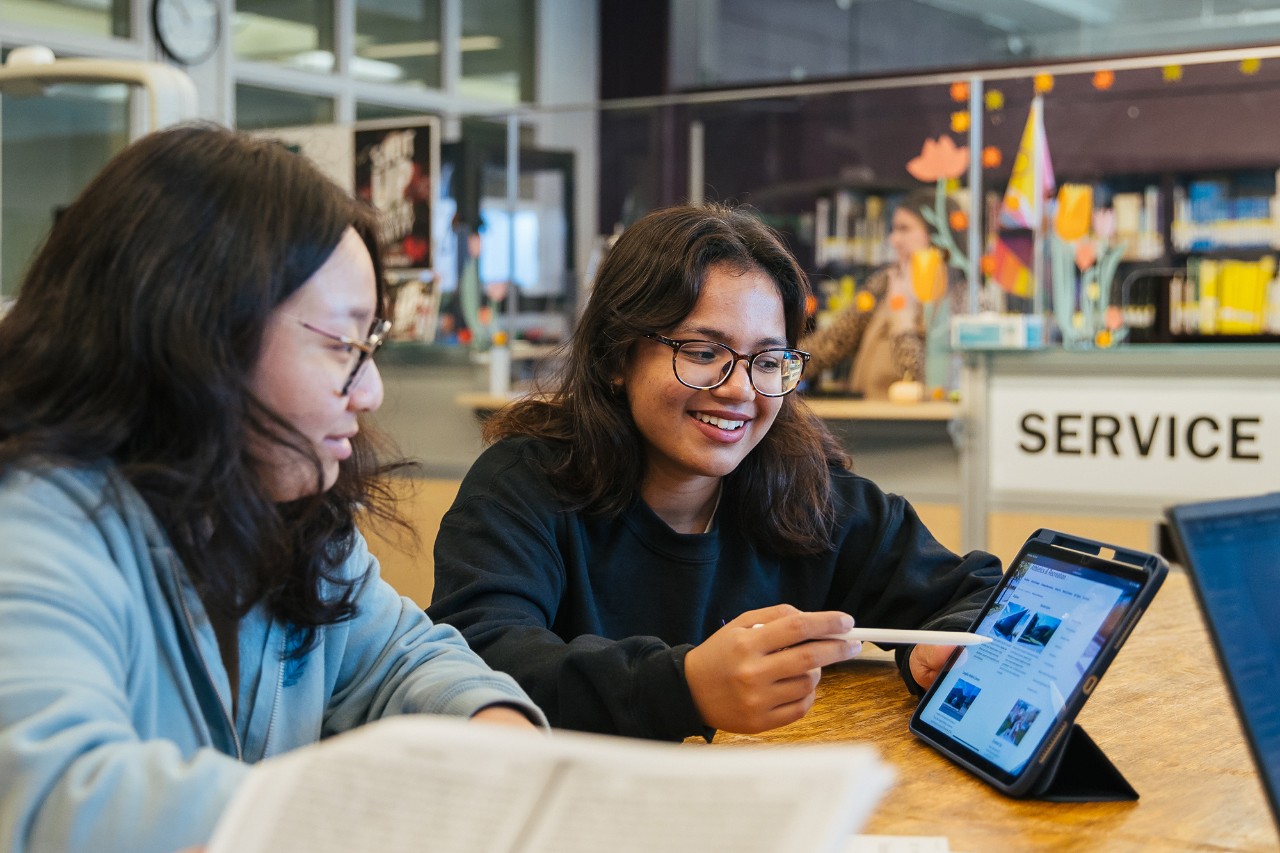 Two female students sitting at a desk. One is using a stylus to point at a tablet.