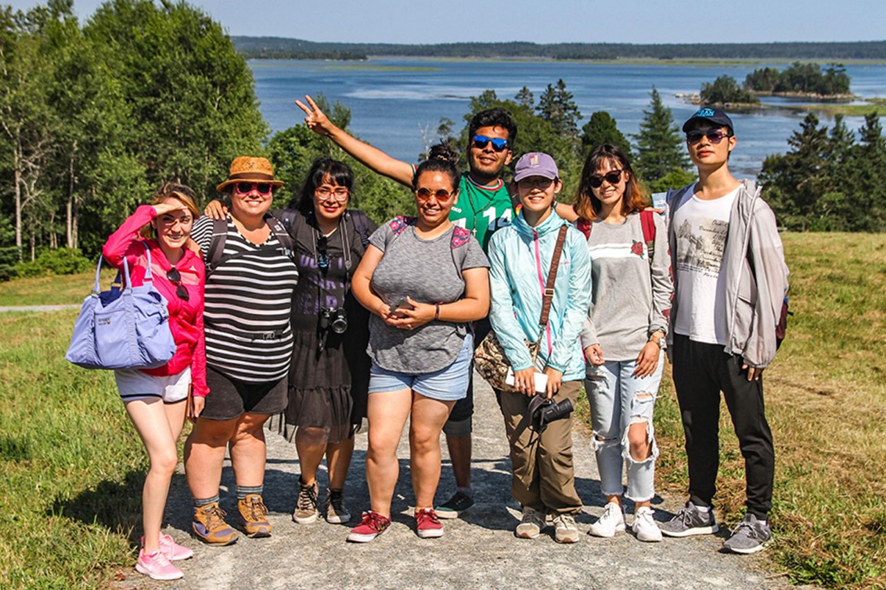 A group of international students wearing sunglasses and smiling into the camera with the ocean and field in the background.